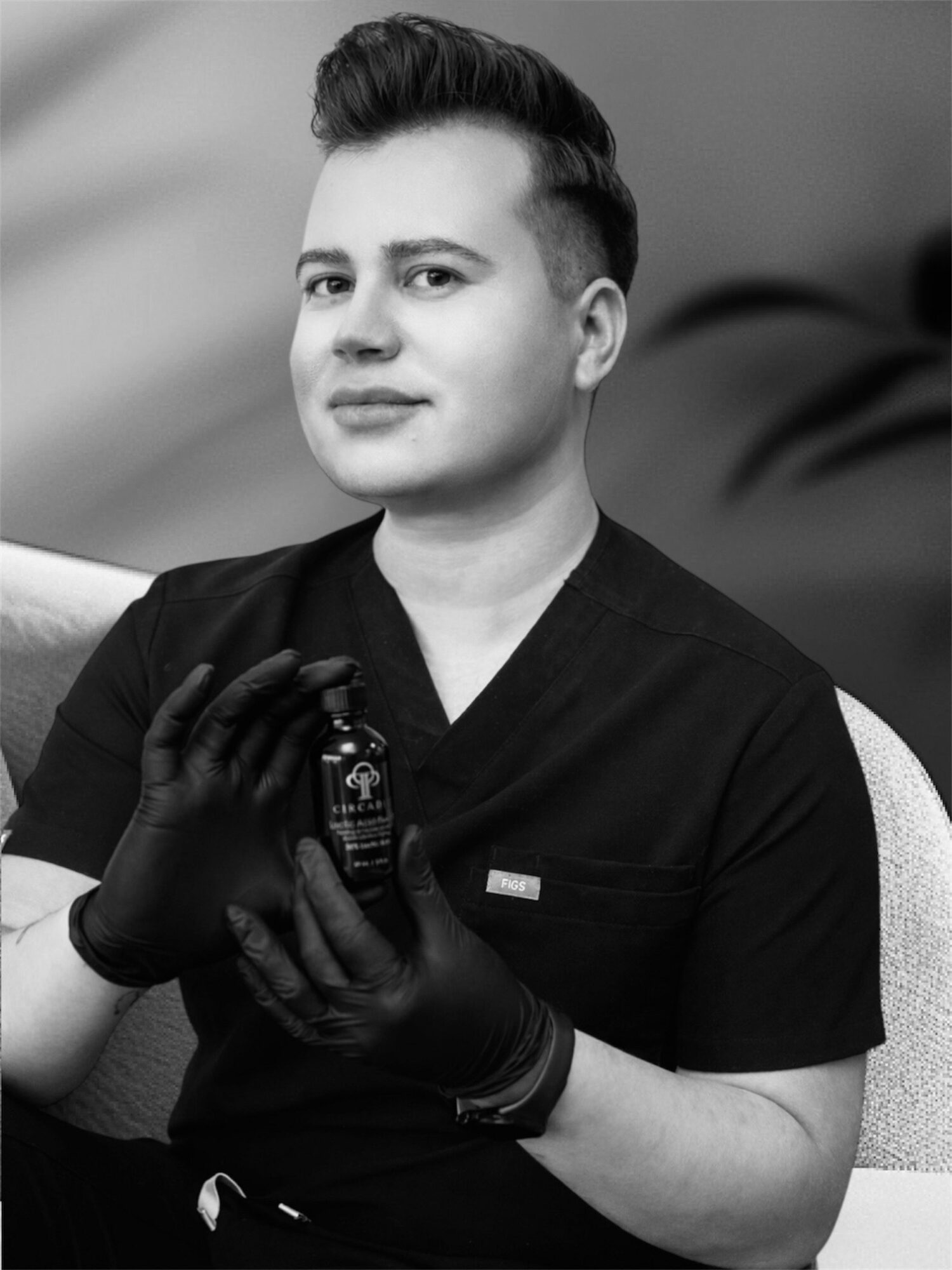 Person in medical scrubs holding a small bottle, wearing gloves, sitting indoors with a blurred background.