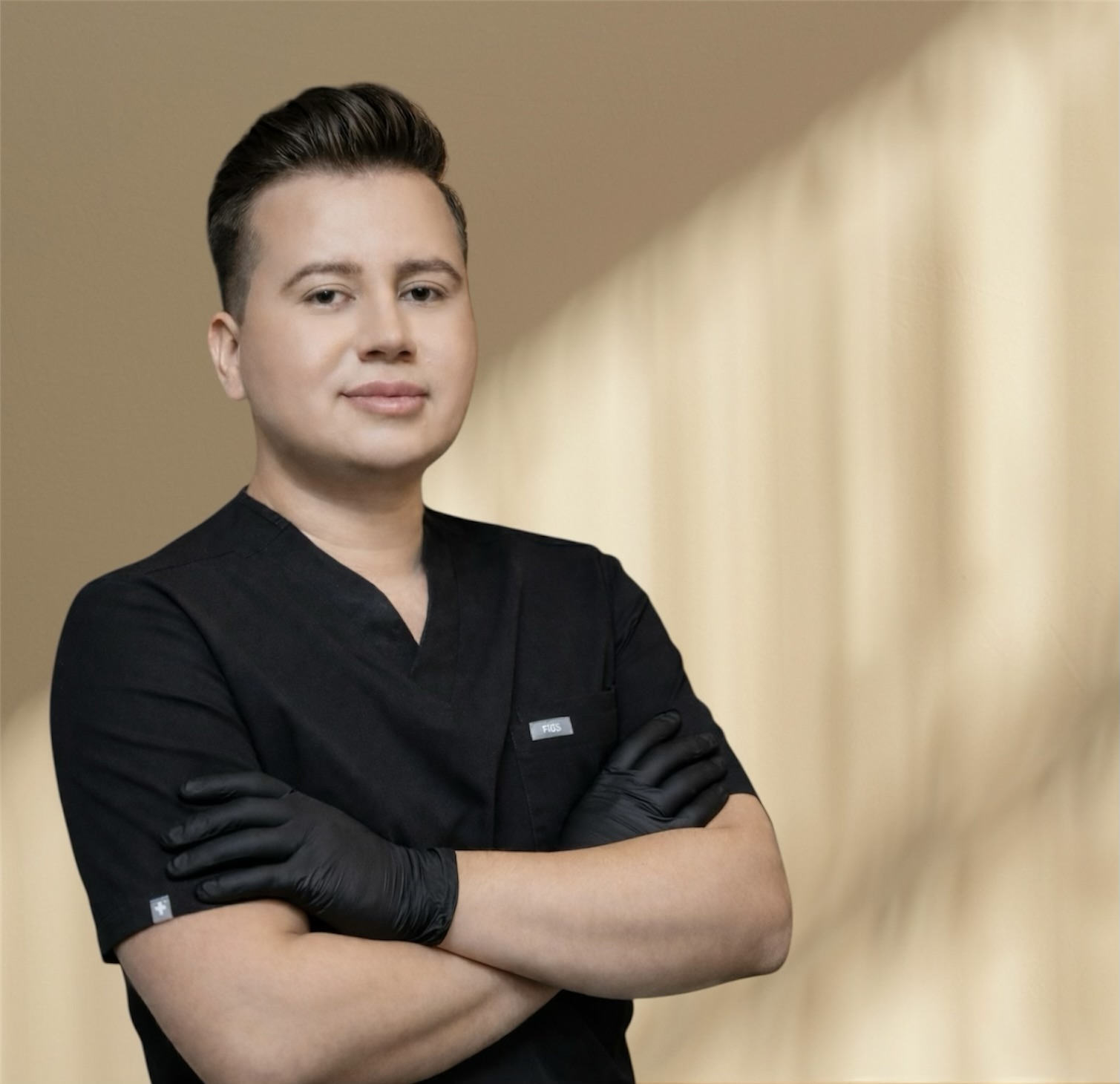 Person with short hair wearing black scrubs and black gloves, arms crossed, standing indoors with beige curtains in background.