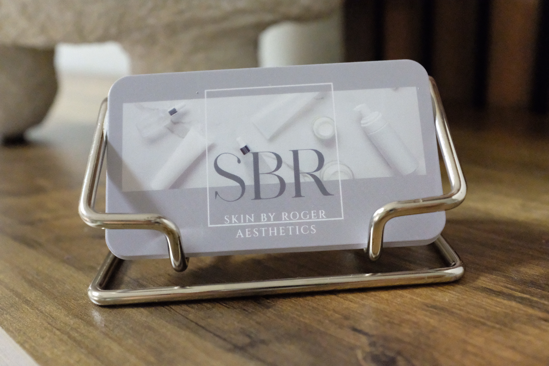 A small rectangular mirror with a metal frame and stand on a wooden surface, reflecting a logo with the text 'SBR' and 'Aesthetics'.