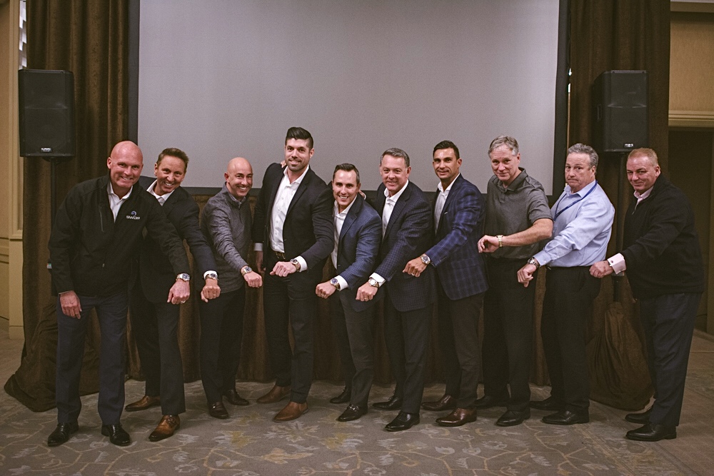 Group of ten men standing in front of a large screen, dressed in formal and semi-formal attire, smiling.
