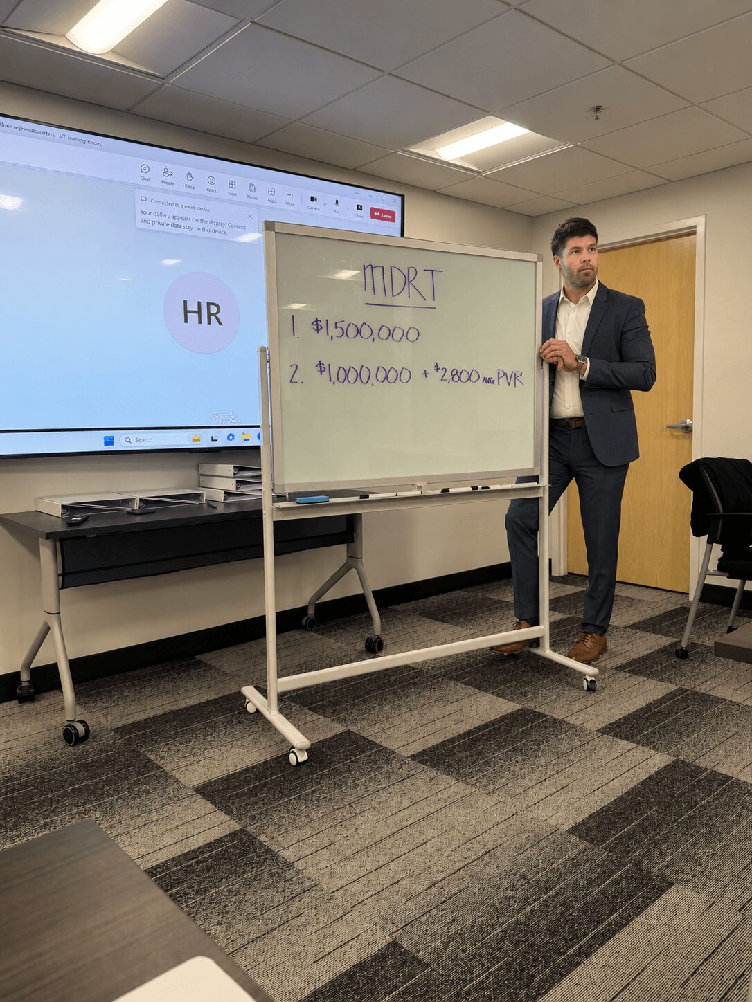 Man in suit standing next to whiteboard with financial calculations, in a conference room with a large screen and carpeted floor.