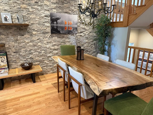Dining room with stone wall, wooden table, six chairs, chandelier, and staircase with wooden railing.