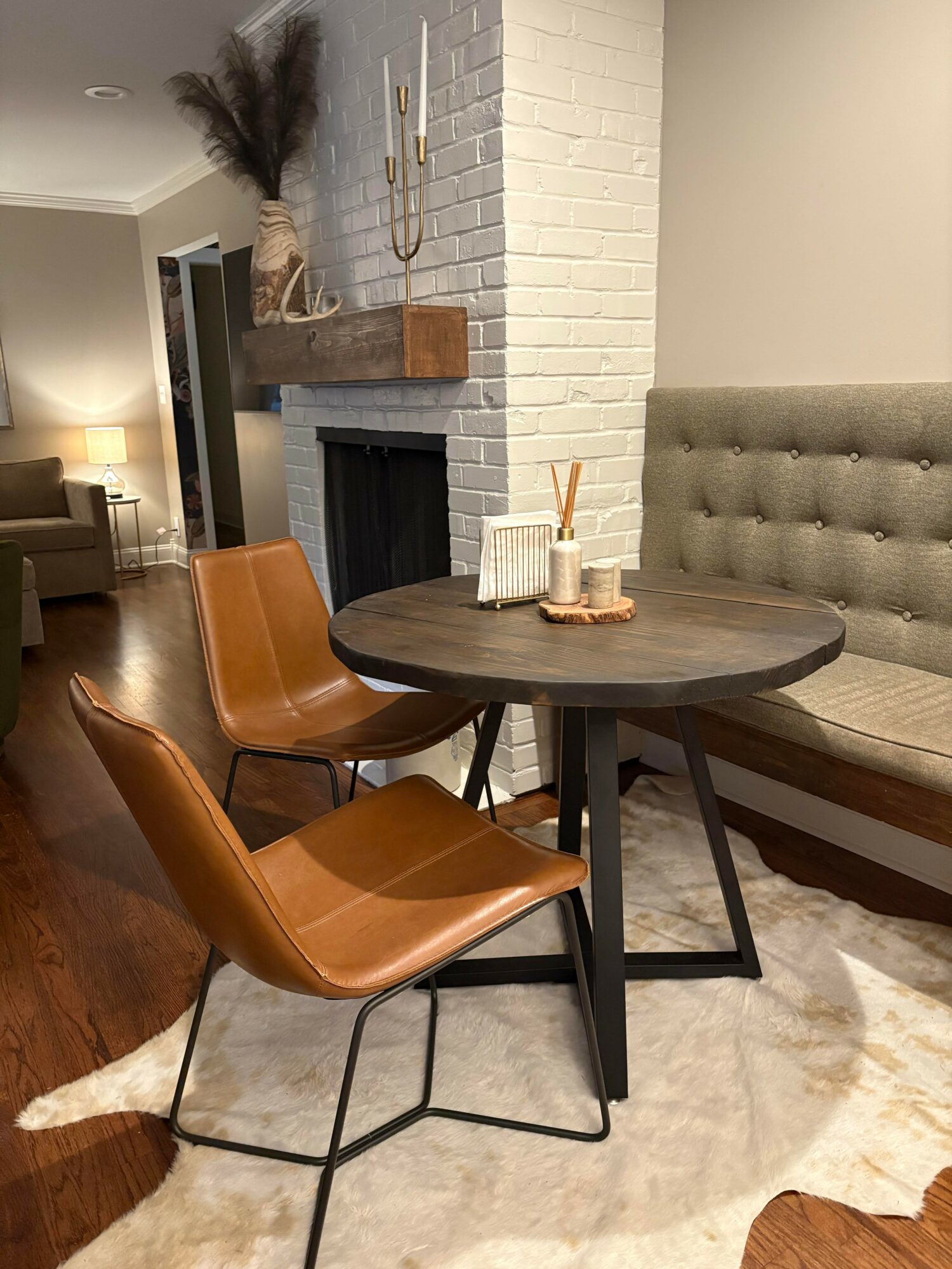 Round wooden table with two brown leather chairs in a living room, fireplace, and beige tufted bench, with a white brick wall.