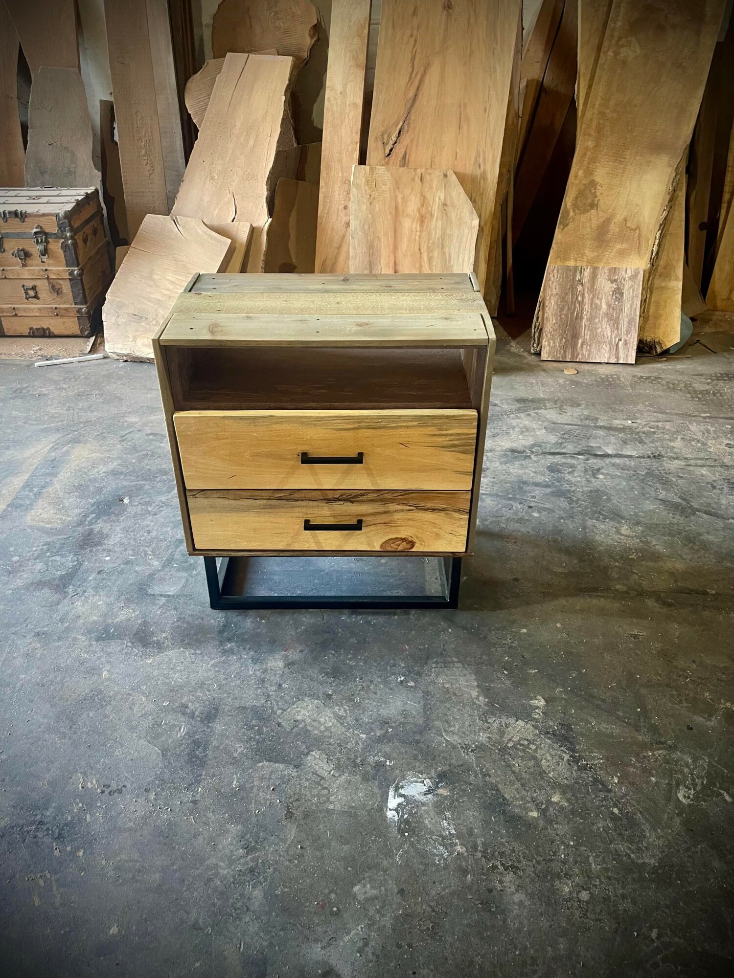 Wooden nightstand with two drawers and black handles, on a concrete floor, surrounded by wood planks and boxes.
