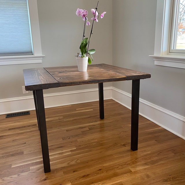 Wooden table with a potted orchid plant on top, in a room with hardwood floor and two windows.