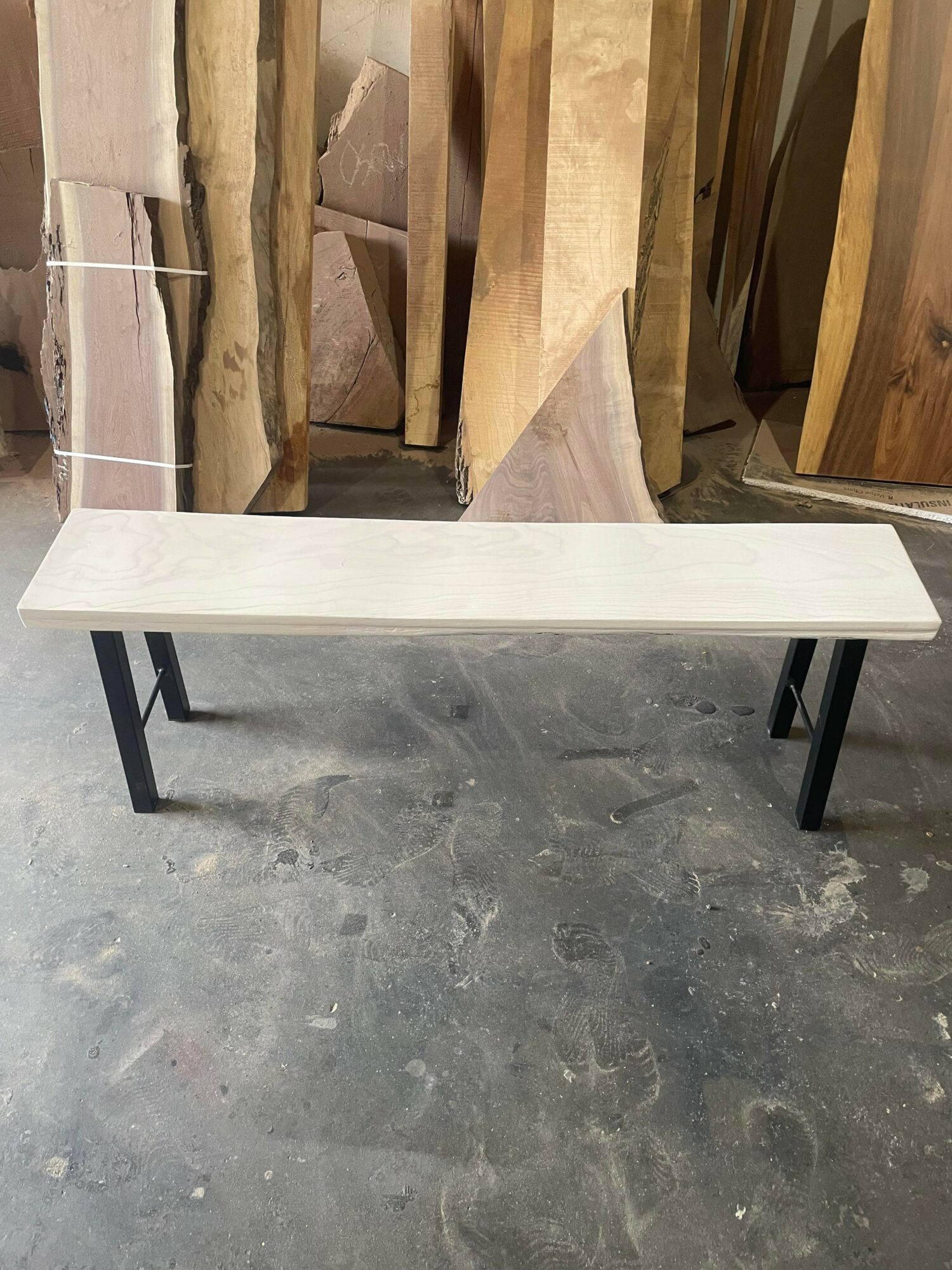Long white bench with black legs in a workshop with wooden slabs in the background.