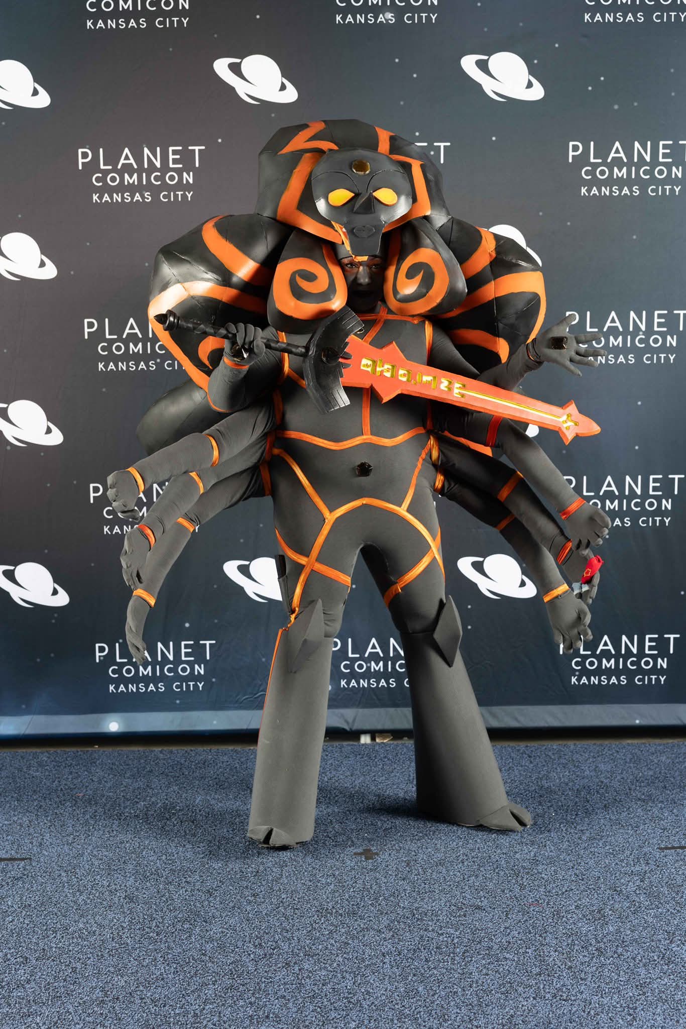 Cosplayer in black and orange armor with a large weapon, standing in front of a Planet Comicon Kansas City backdrop.