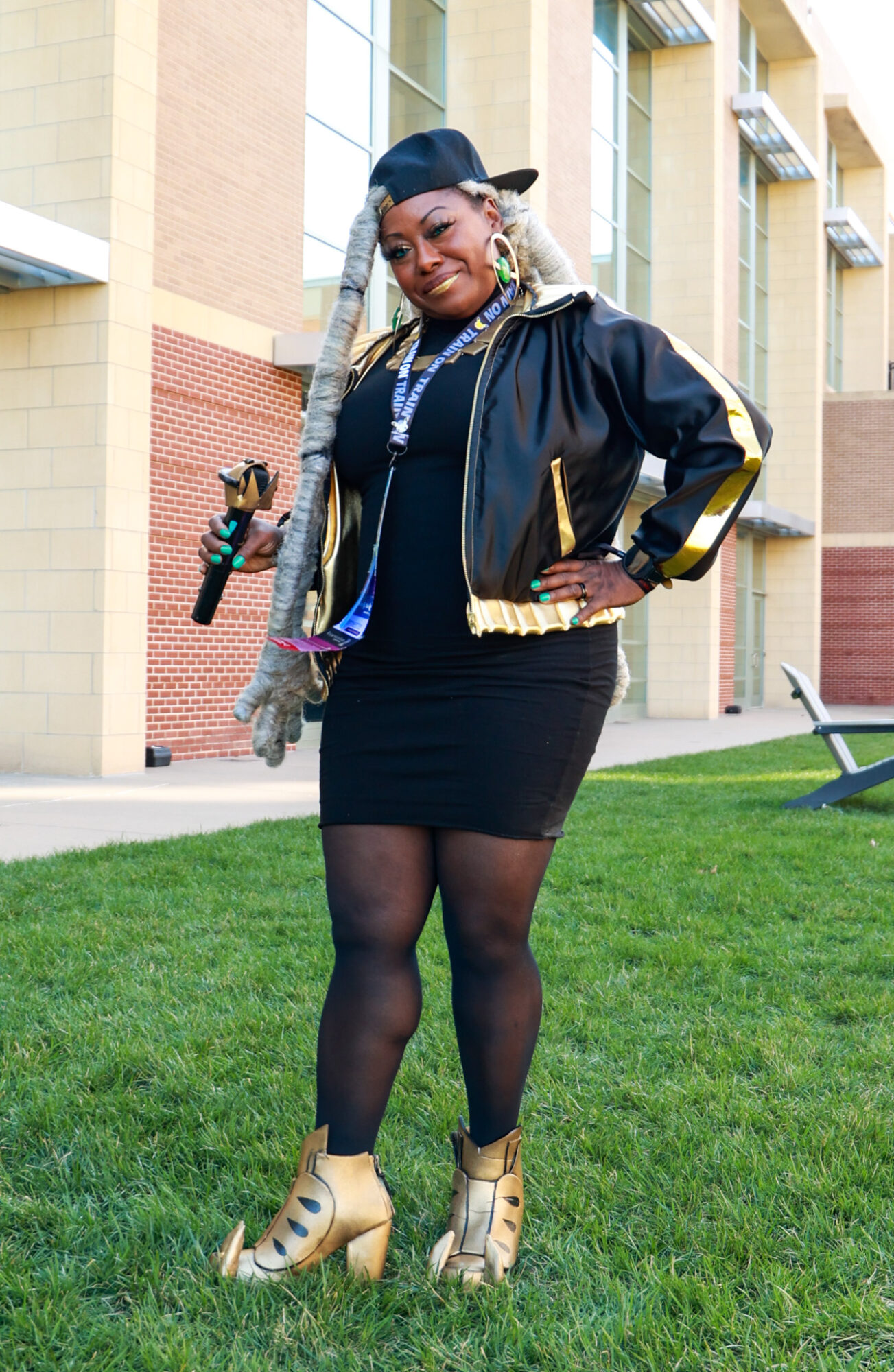 Woman with blonde braids in a black cap, black jacket with gold accents, black dress, and beige high-heeled shoes, standing on grass in front of a building.
