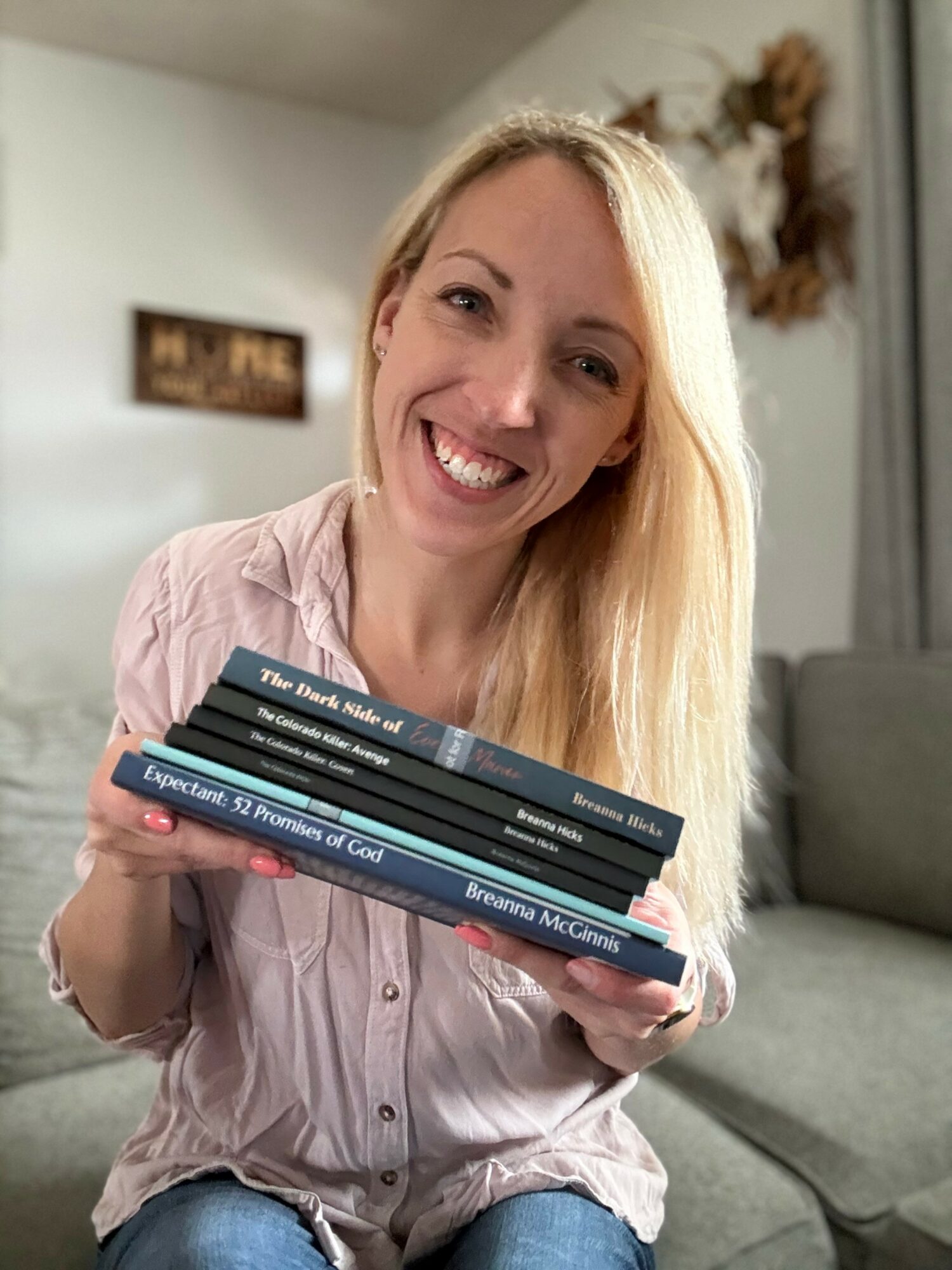 Woman with long blonde hair smiling and holding a stack of books indoors.