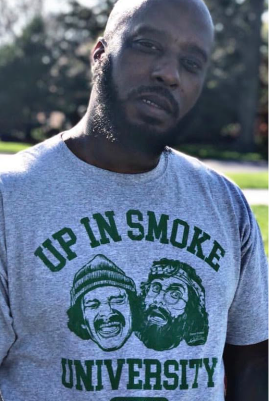 Man wearing a gray T-shirt with green text and graphics outdoors, trees in background.