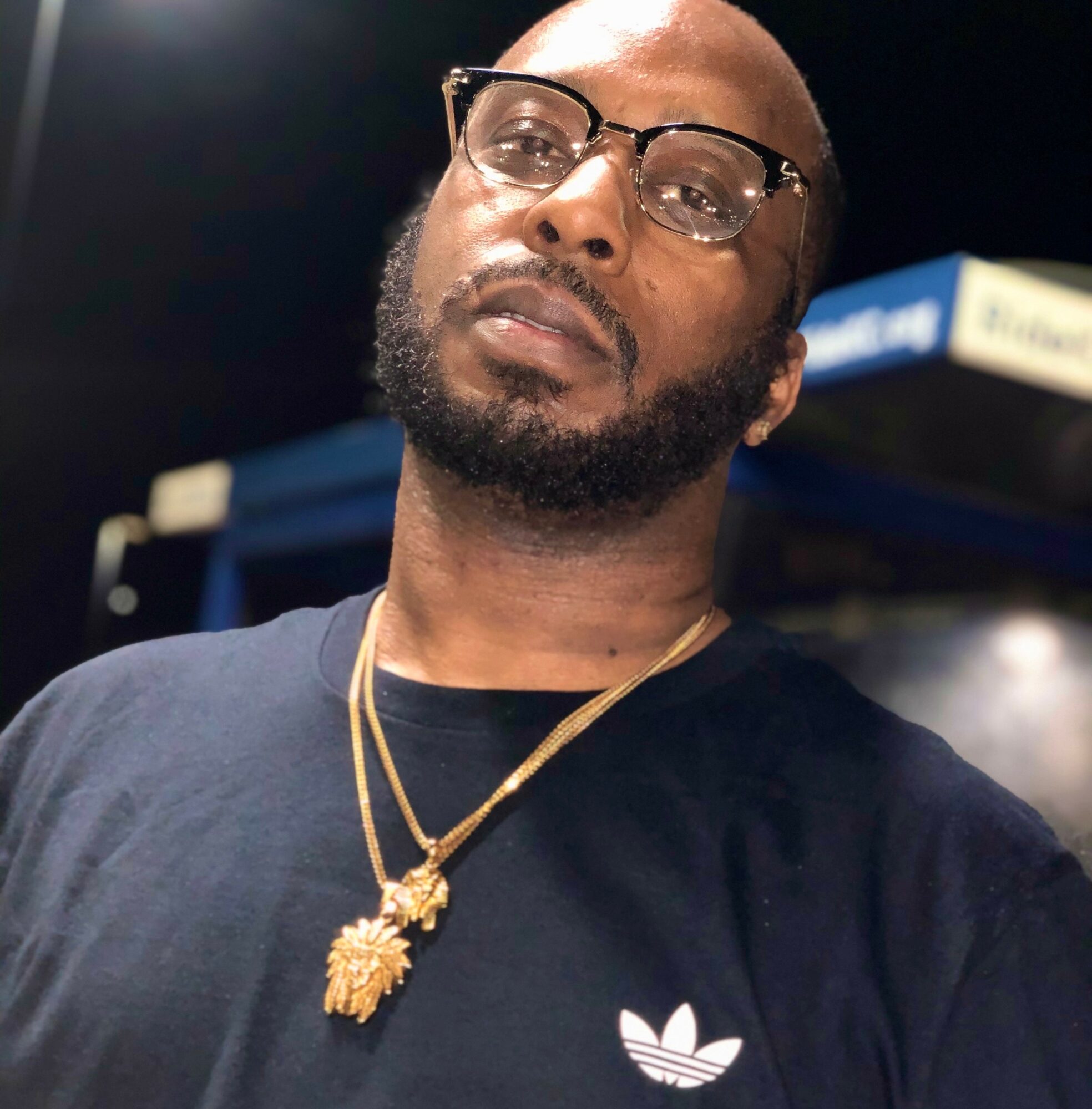 Man with glasses, beard, wearing a black shirt and gold necklaces, looking at the camera.