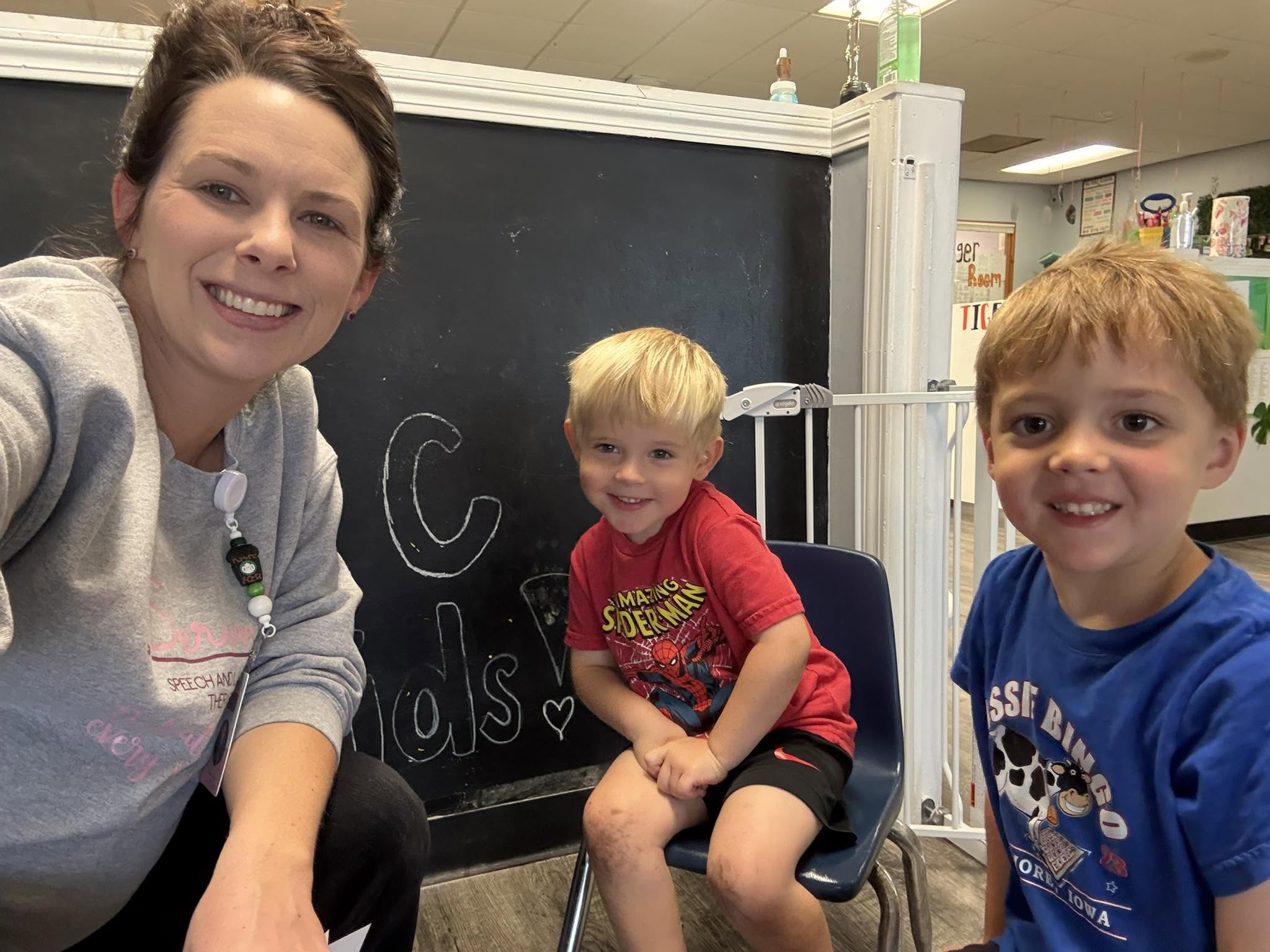 Woman and two young boys smiling in a classroom with a blackboard behind them.