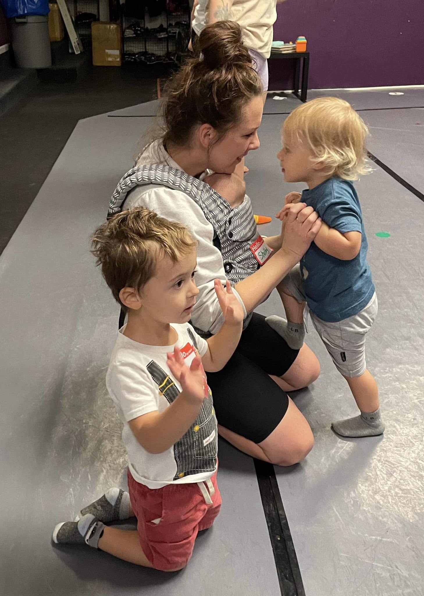 Kneeling woman interacts with two young children on a gray floor, one child standing and the other sitting, in an indoor setting.