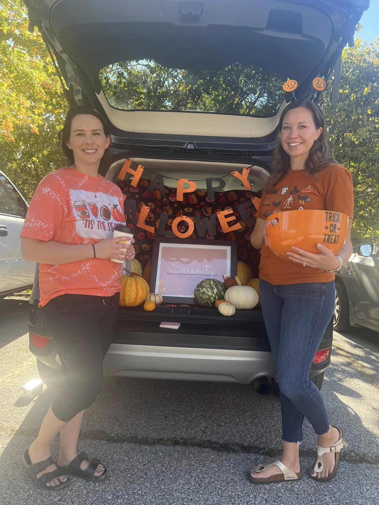 Two women stand outdoors behind a vehicle with a Halloween display, holding pumpkins and a tablet, with a 'Happy Halloween' banner.