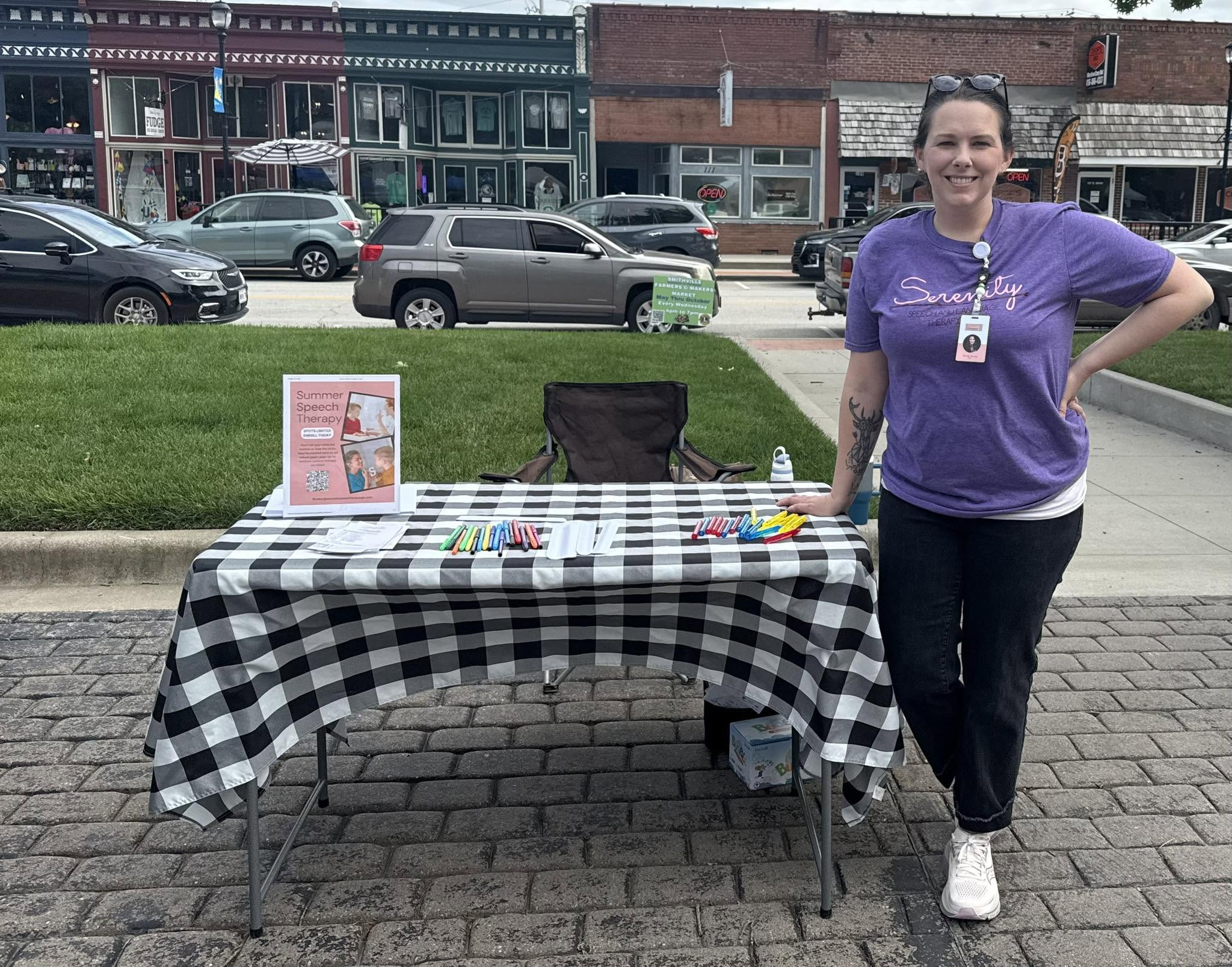 Woman standing next to a table with items, in an outdoor urban setting with parked cars and buildings behind.