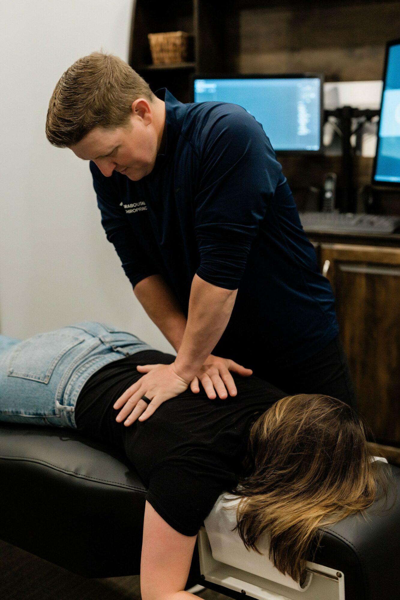 Person performing chiropractic adjustment on a patient lying face down on a table, in a clinical setting with monitors in the background.