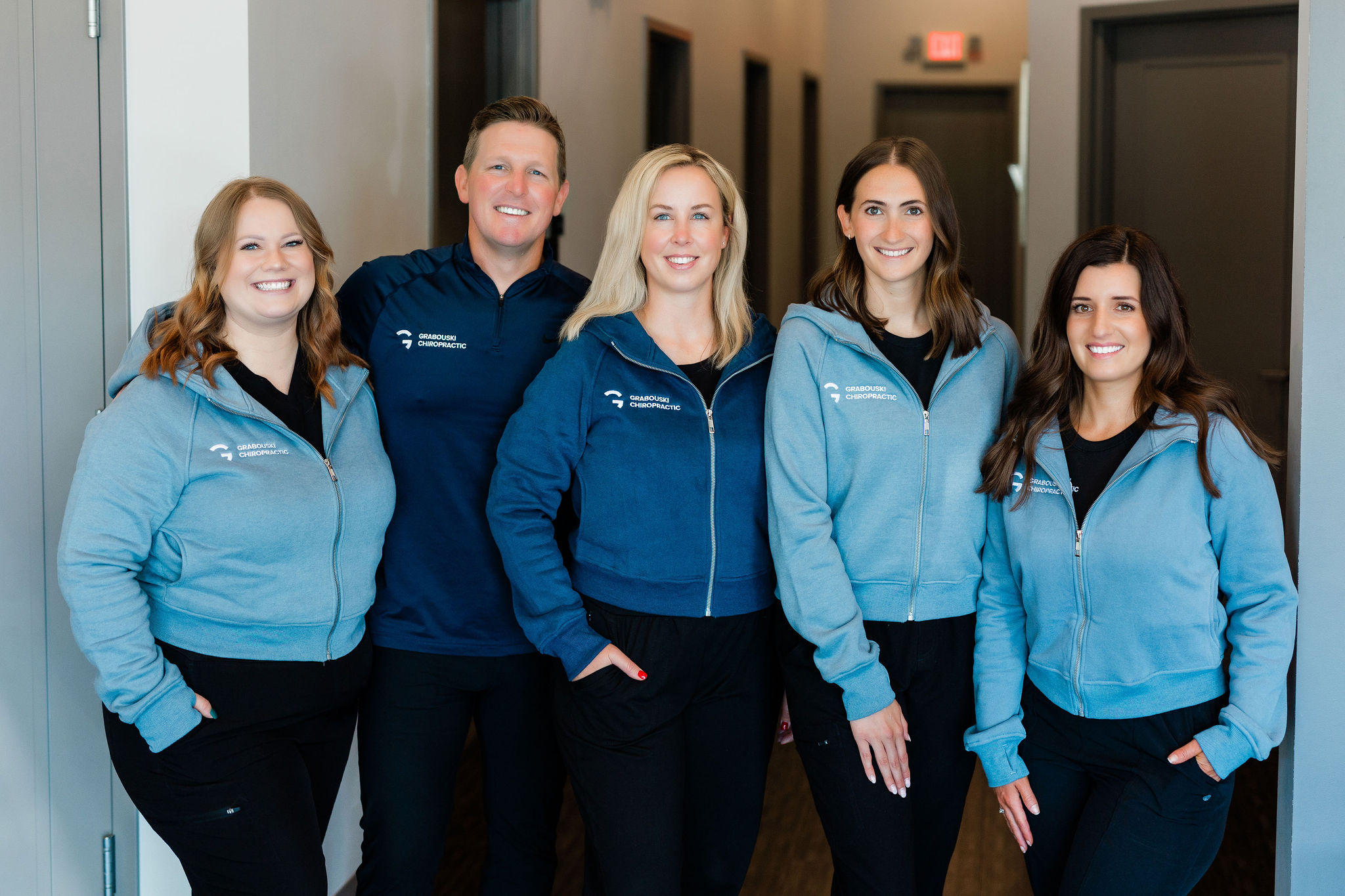 Group of five smiling people standing indoors, wearing blue jackets, in a hallway with gray walls and doors.