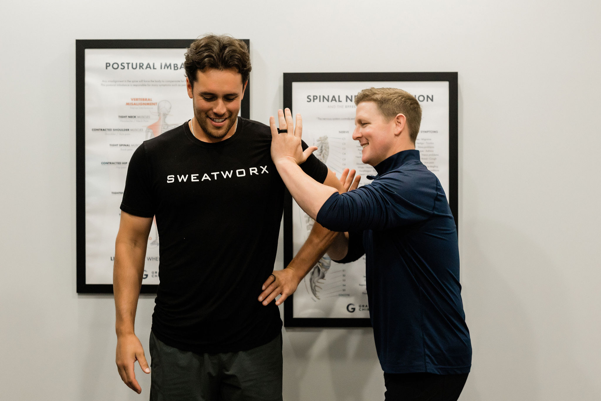 Two men smiling and high-fiving in front of framed posters on a white wall.