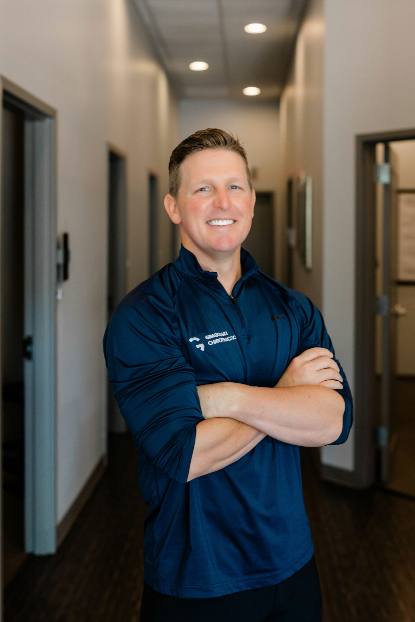 Man with short hair smiling, arms crossed, standing in a hallway with doors on both sides.