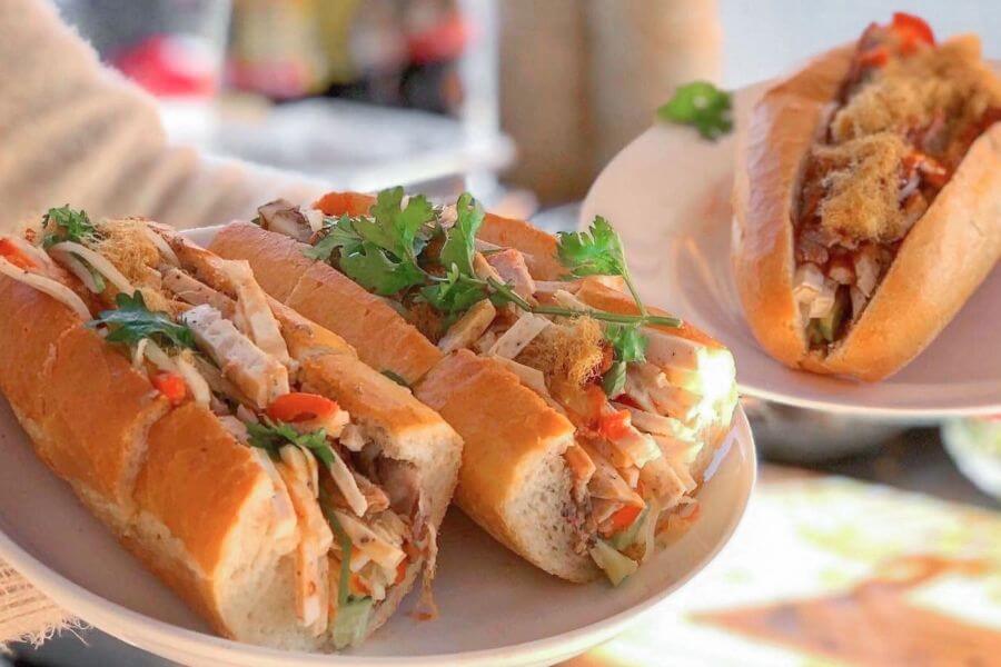 Two sandwiches with shredded meat, vegetables, and cilantro on a white plate, one partially eaten, in a casual setting.