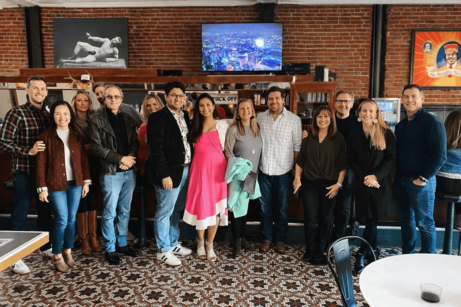 Group of 12 people standing in a room with brick wall, TV, and artwork, smiling for a photo.