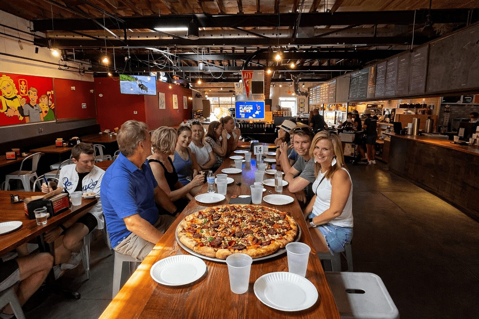Group of people sitting at a long table in a restaurant with a large pizza in the center, smiling and talking.