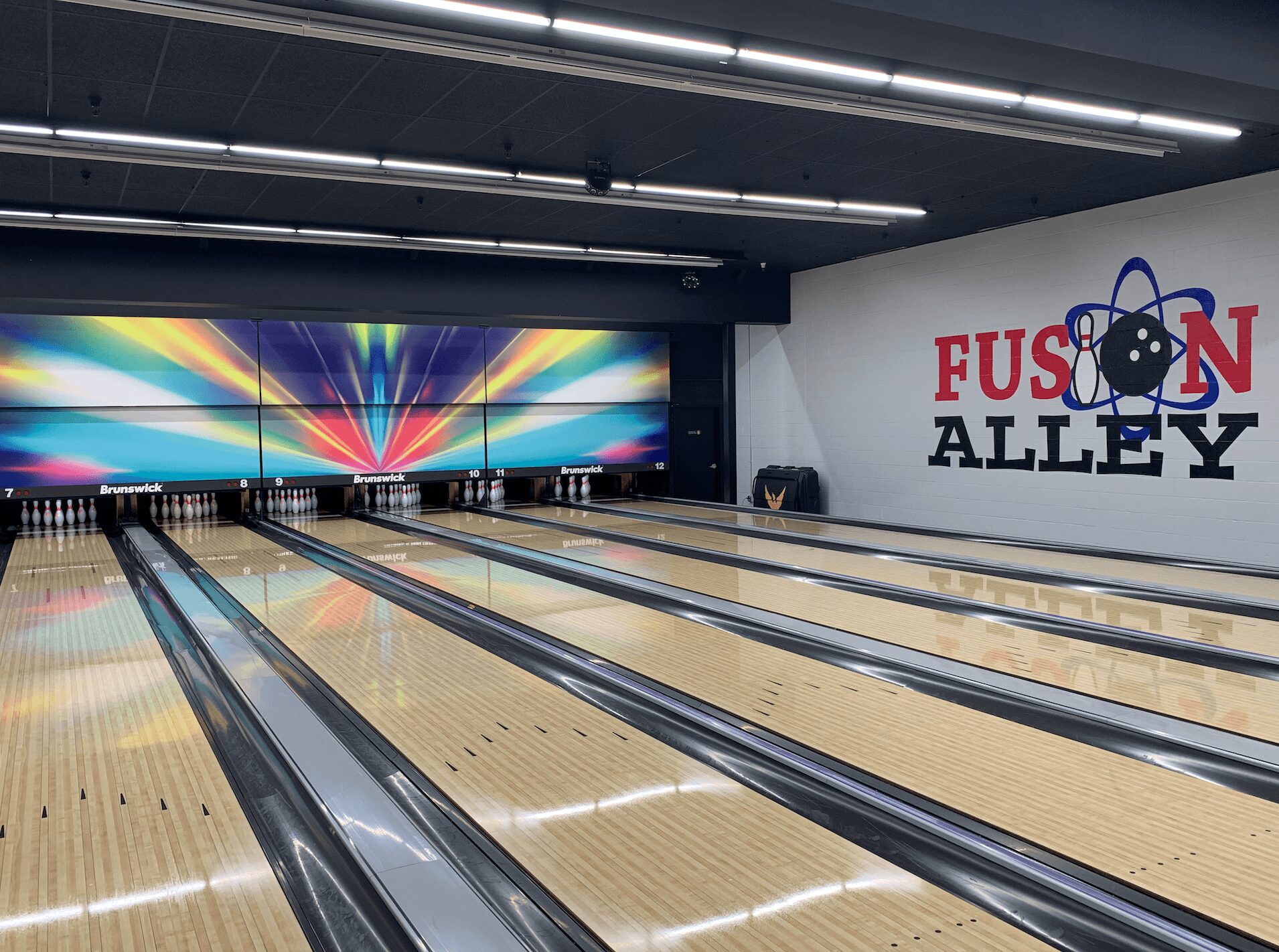 Bowling lanes with colorful digital display and a wall with 'FUSION ALLEY' logo, ceiling lights, and seating area.