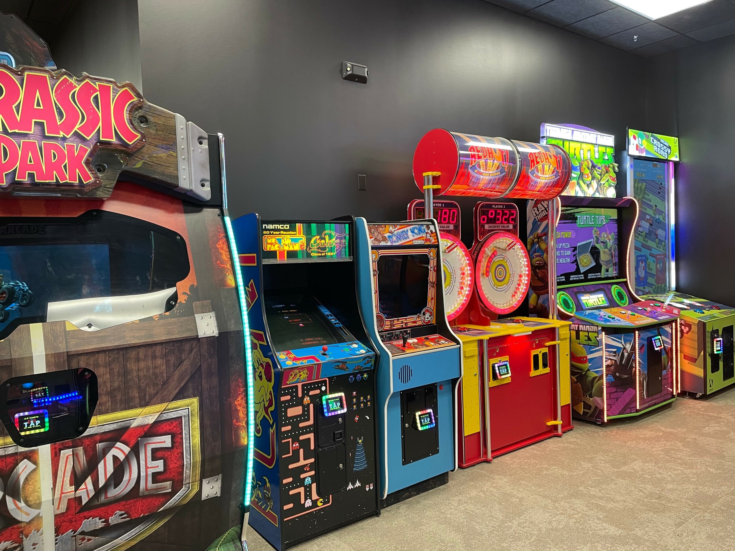 Row of arcade game machines in a gaming center, with colorful screens and controls, against a dark wall.