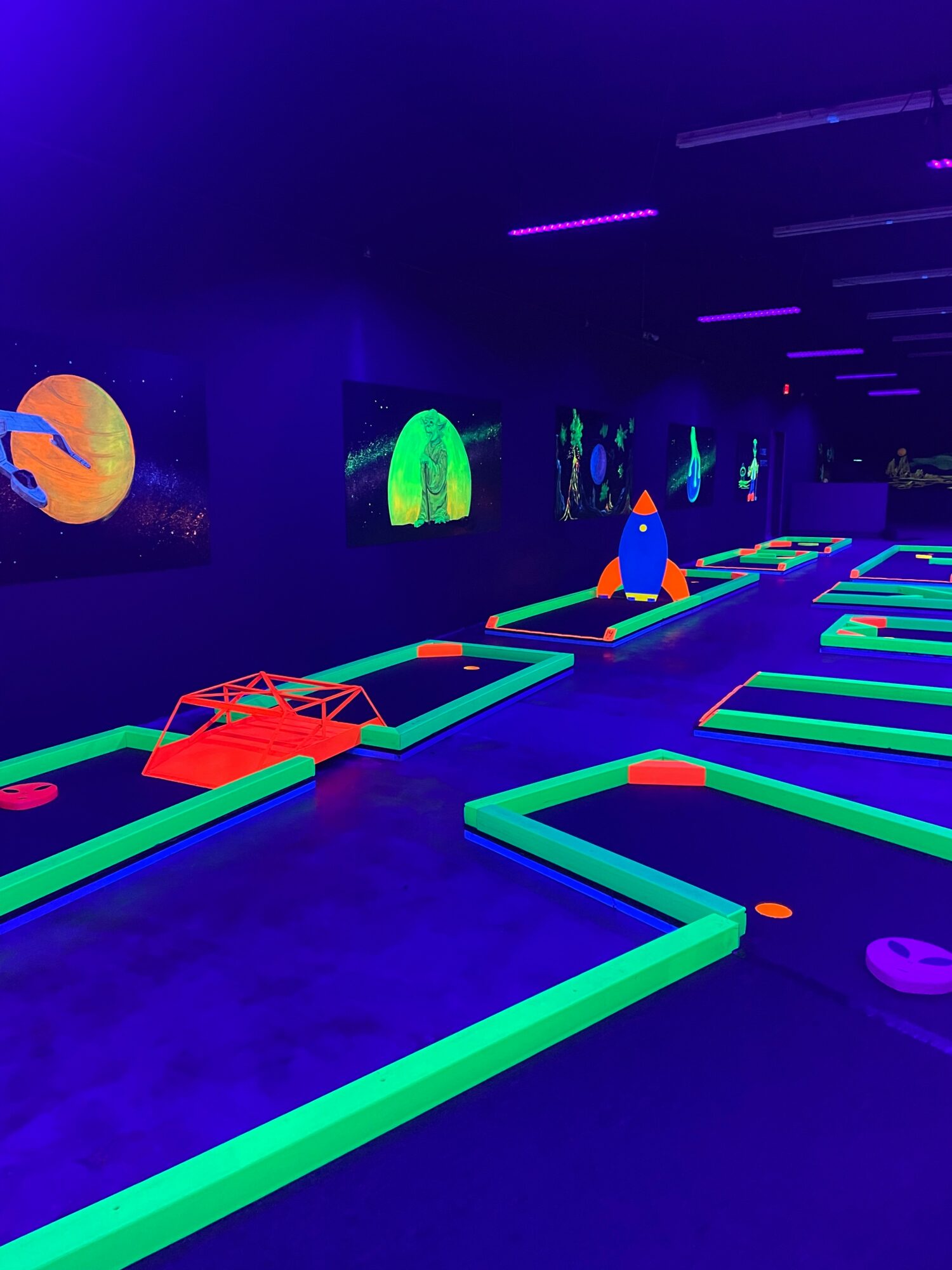 Glow-in-the-dark mini golf course with neon-lit obstacles and screens, illuminated in dark room, colorful and futuristic atmosphere.