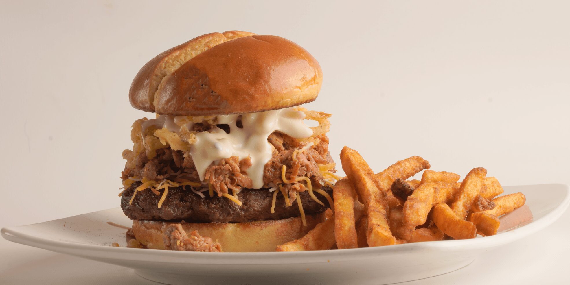 Burger with fried chicken, lettuce, and sauce, served with curly fries on a white plate against a plain background.