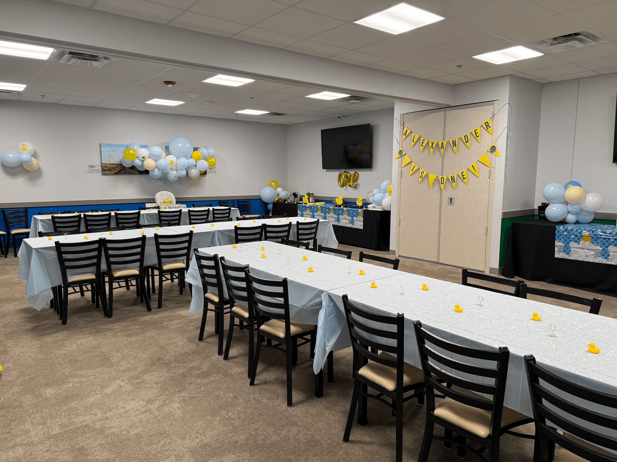 Party room decorated with blue, white, and yellow balloons, with long tables covered in light blue tablecloths and black chairs.