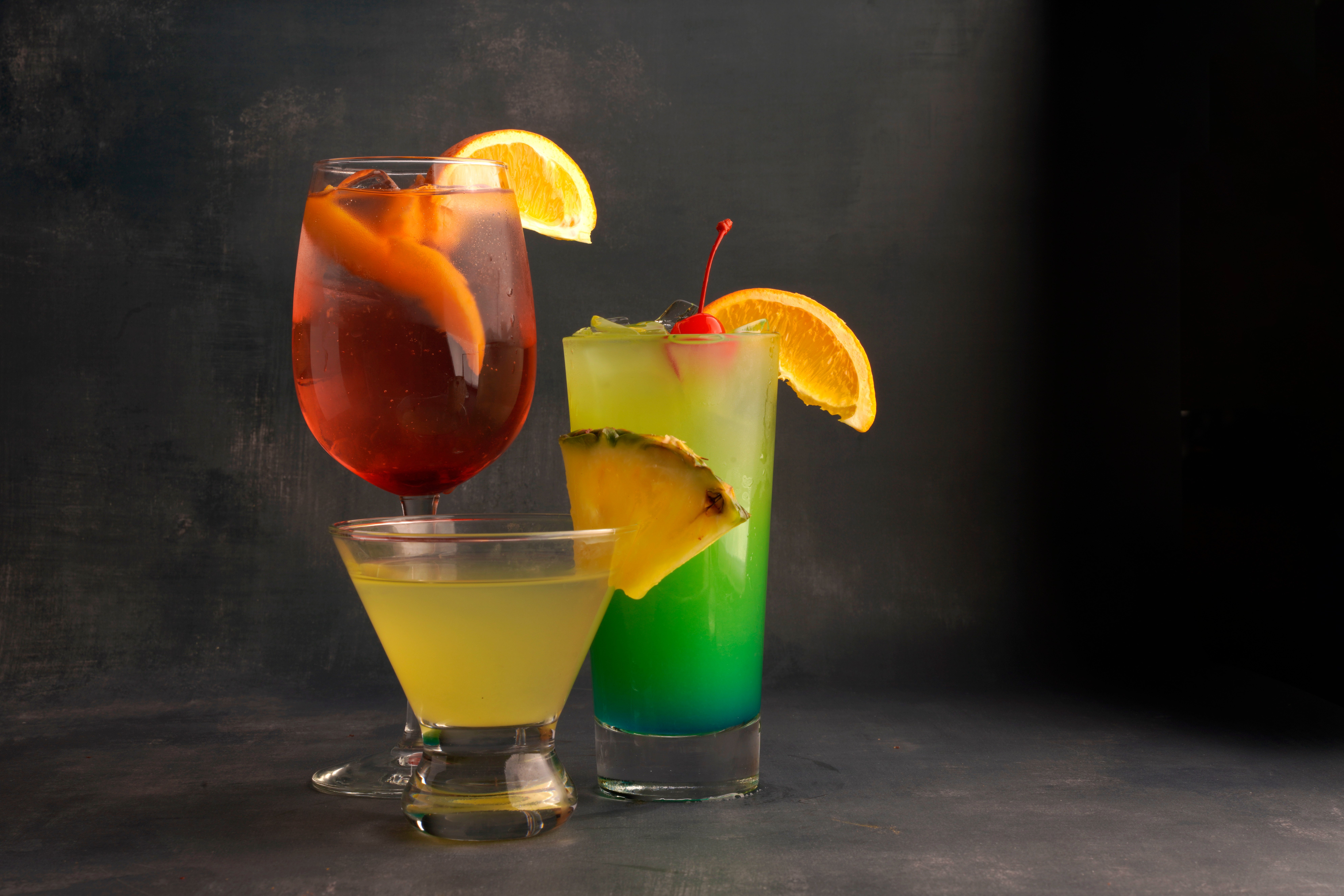 Three colorful cocktails with fruit garnishes on a dark surface, one in a glass with a lemon slice, one with pineapple and cherry, and a yellow drink in a short glass.