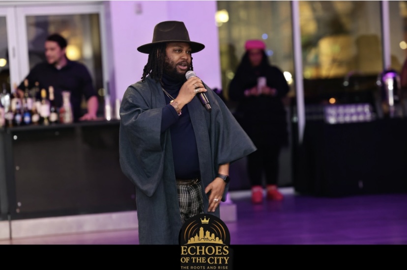 Man speaking into microphone at event, with bar and people in background, indoors with purple lighting.
