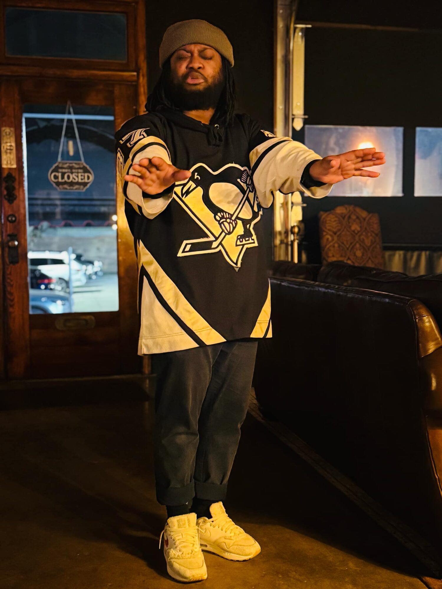 Person with beard and beanie in a hockey jersey, standing indoors with arms extended, near a window and door with a 'Closed' sign.