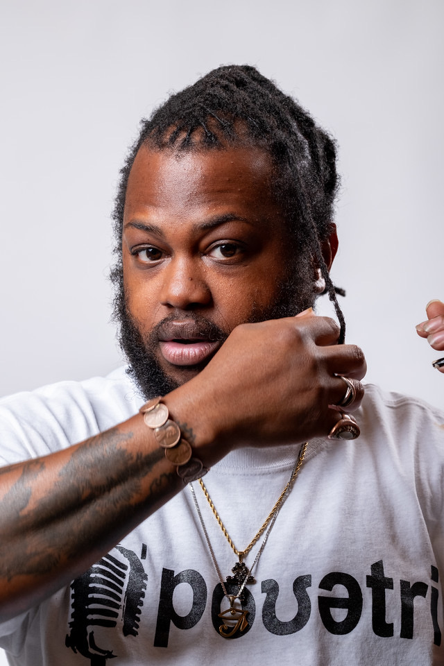 Man with dreadlocks and beard adjusting his shirt, wearing a white t-shirt and jewelry, against a plain background.