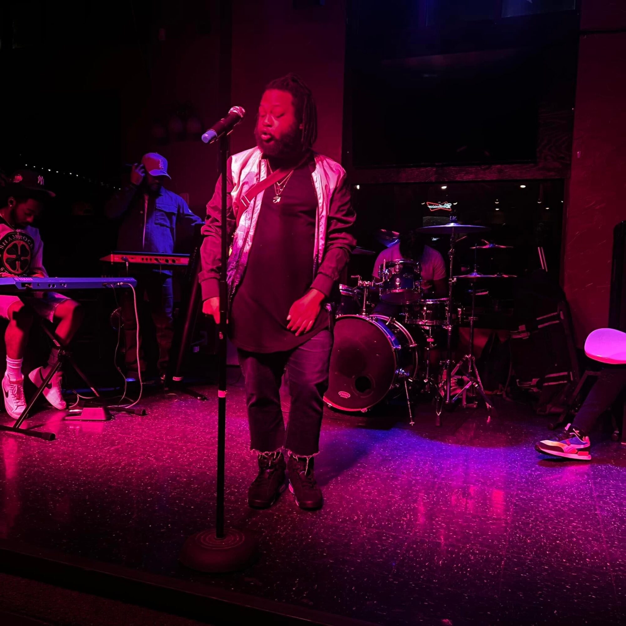 Person singing into a microphone on stage with band members and instruments, illuminated by pink and purple stage lights.