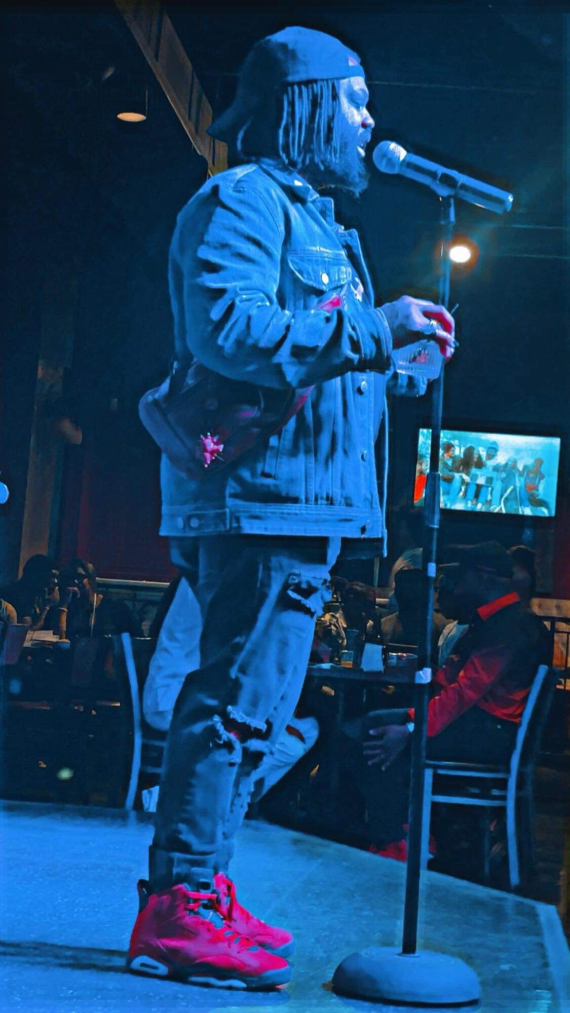Person with long hair and hat singing into a microphone on stage, wearing a denim jacket and red shoes.