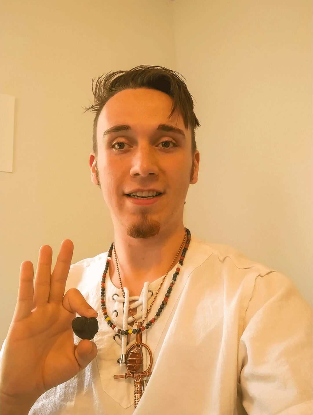 Young man with short dark hair and a goatee, smiling, making an OK gesture with his right hand, wearing layered necklaces, in a light-colored room.