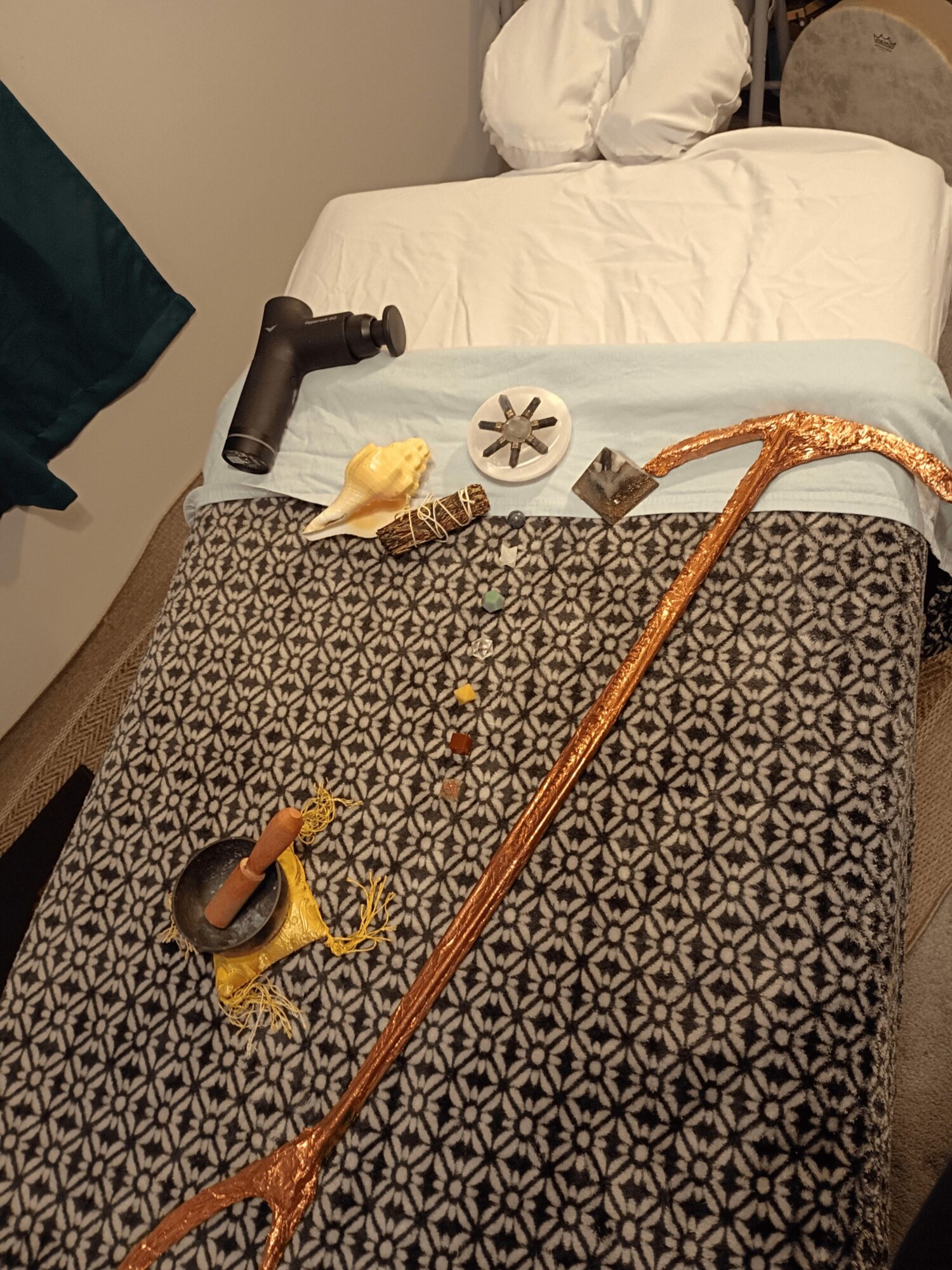 Objects on a patterned bedspread include a wooden staff, a shell, a small dish, and a yellow cloth, with a white pillow in the background.