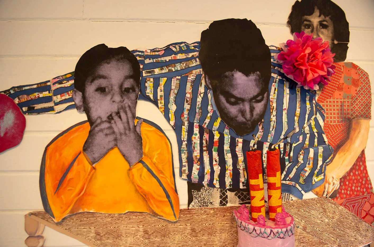 Three people sitting at a table with a birthday cake and candles, one person wearing a bright orange shirt, another with a striped shirt, and a third with a floral dress holding a large pink flower. The image appears to be in black and white with some color accents.