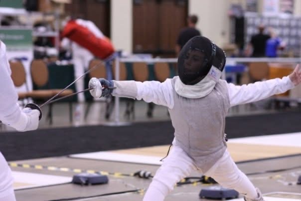 Fencer in white protective gear extends sword in a fencing match, with a blurred background of tables and people.