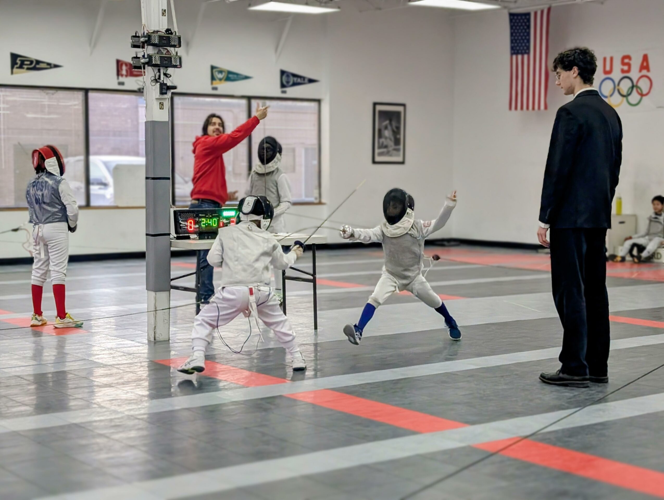 Children fencing in a gym with an instructor, American flag, and Olympic flags on the wall.