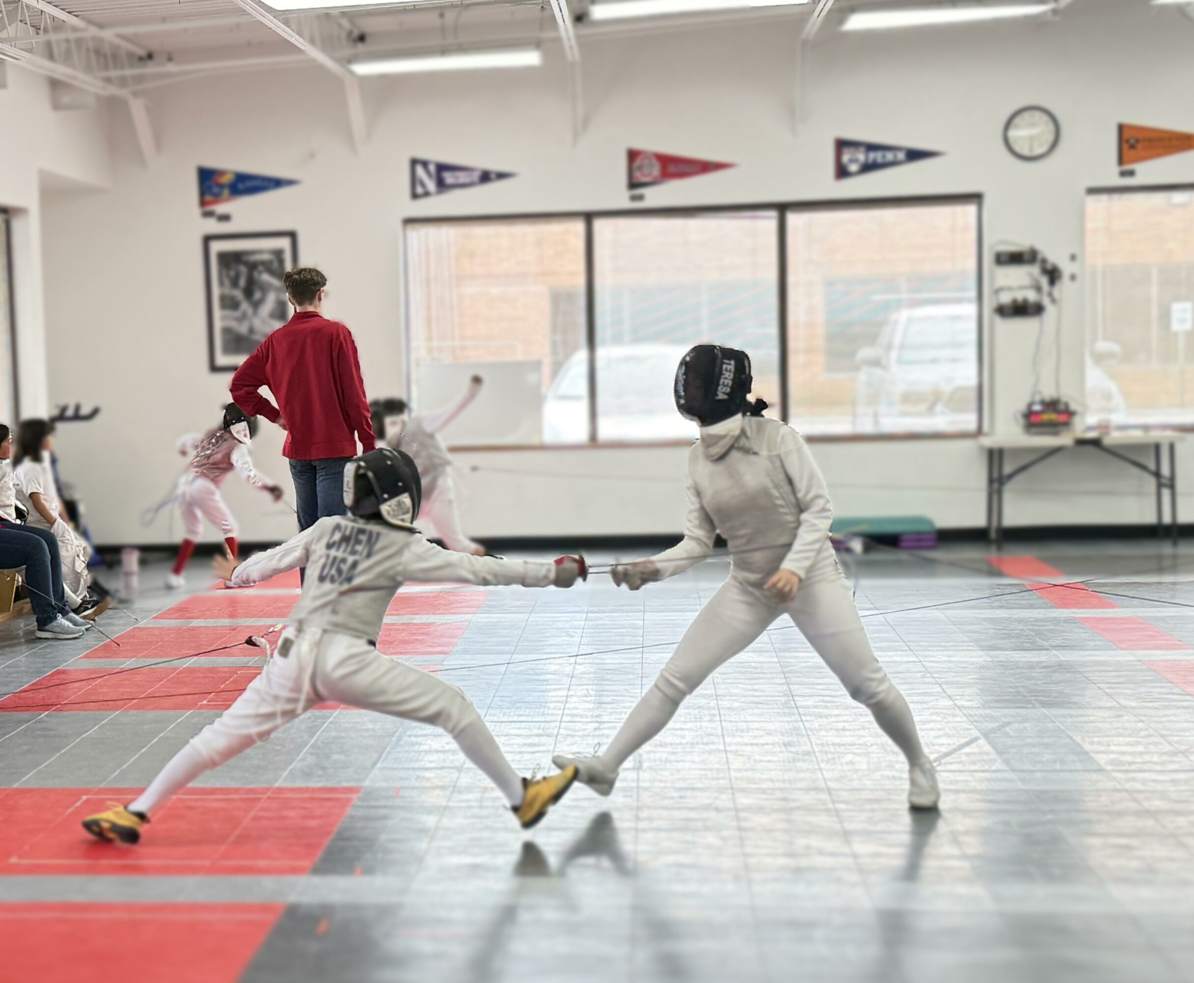 Two young girls in fencing gear sparring in a gym, holding swords and reaching out to each other, with other children and an adult in the background.