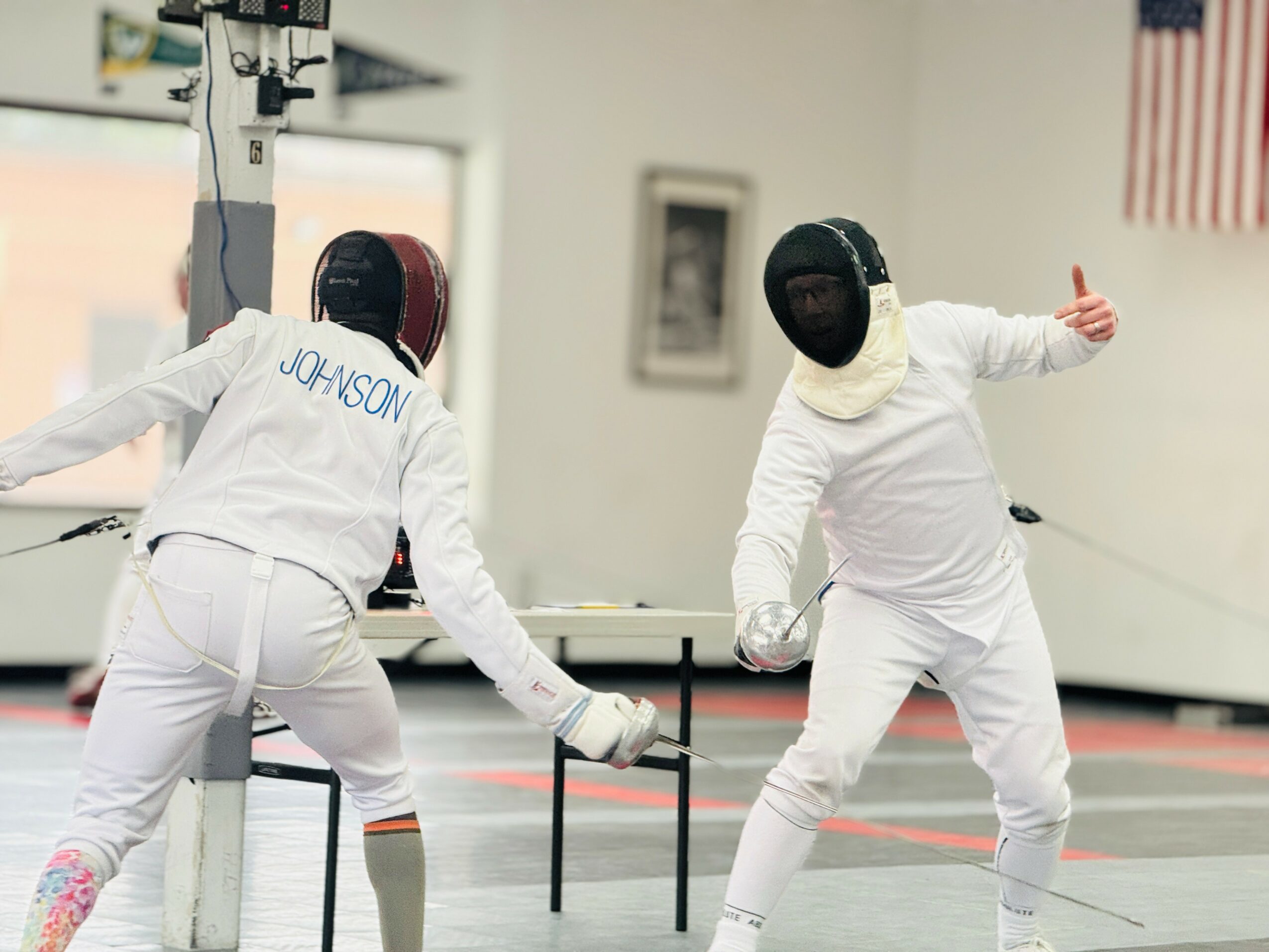 Two fencers in white uniforms and masks practicing in a gym with an American flag on the wall.