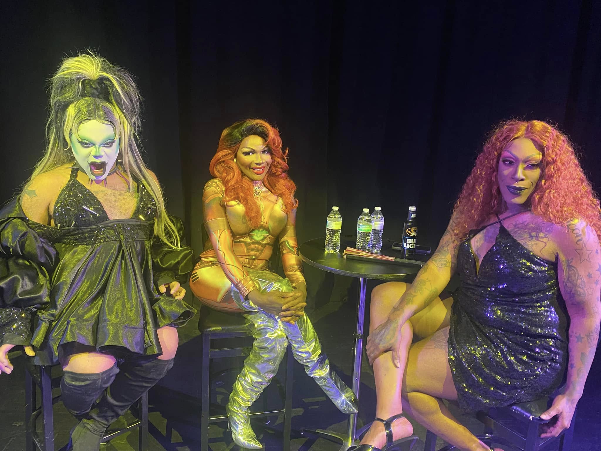 Three performers with colorful costumes and makeup on stage, two sitting and one standing, with water bottles on a table.