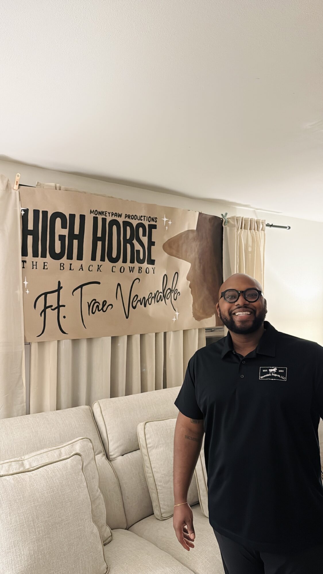 Man with glasses and beard smiling, standing in front of a beige couch and a banner that reads 'High Horse' and 'Ft. Tare Vannell'.