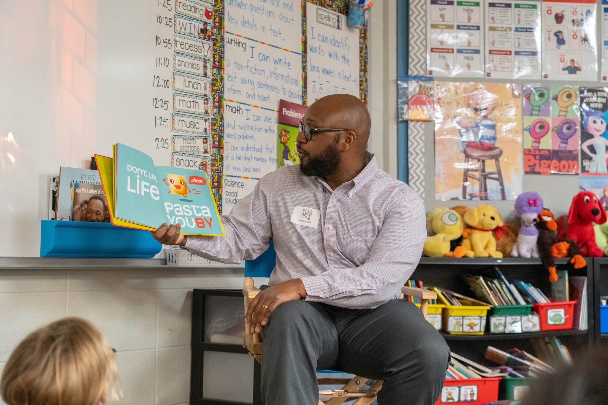 Adult man sitting in a classroom with children, colorful books, and toys on shelves behind him.