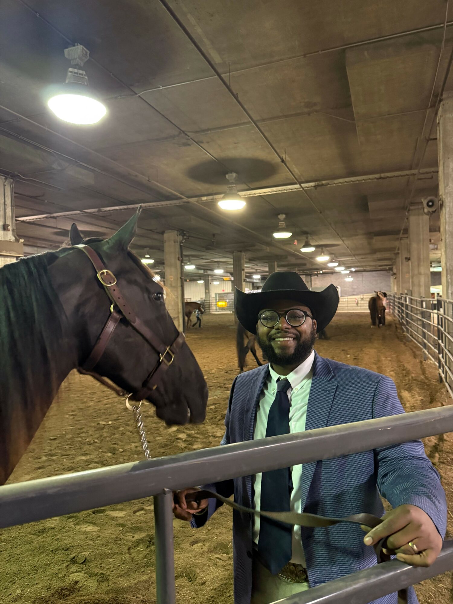 Person wearing glasses, a hat, and a blue jacket standing outdoors near a metal railing, holding a leash, with a horse nearby.