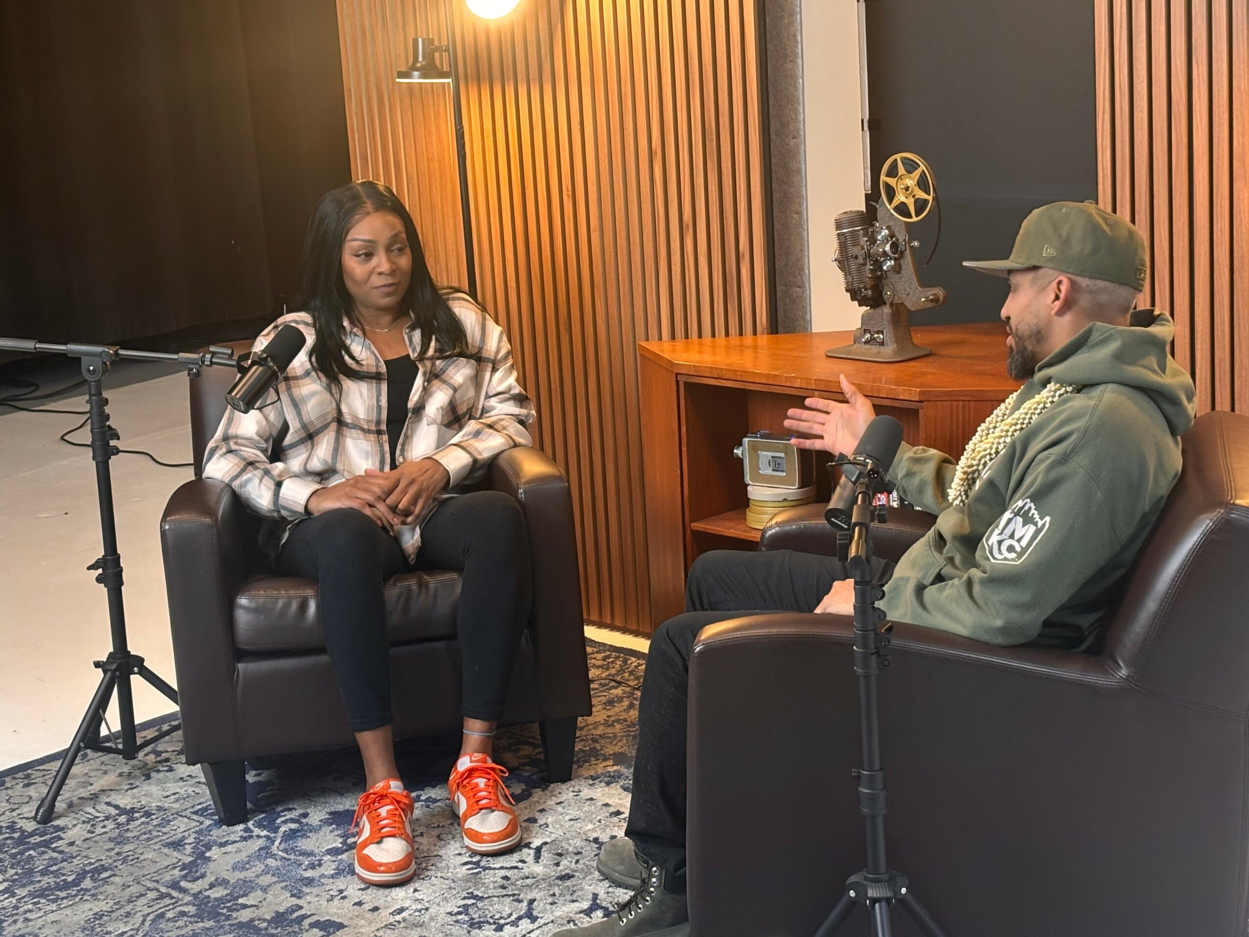 Two people sit in a recording studio, engaged in conversation, with microphones and soundproof walls visible.