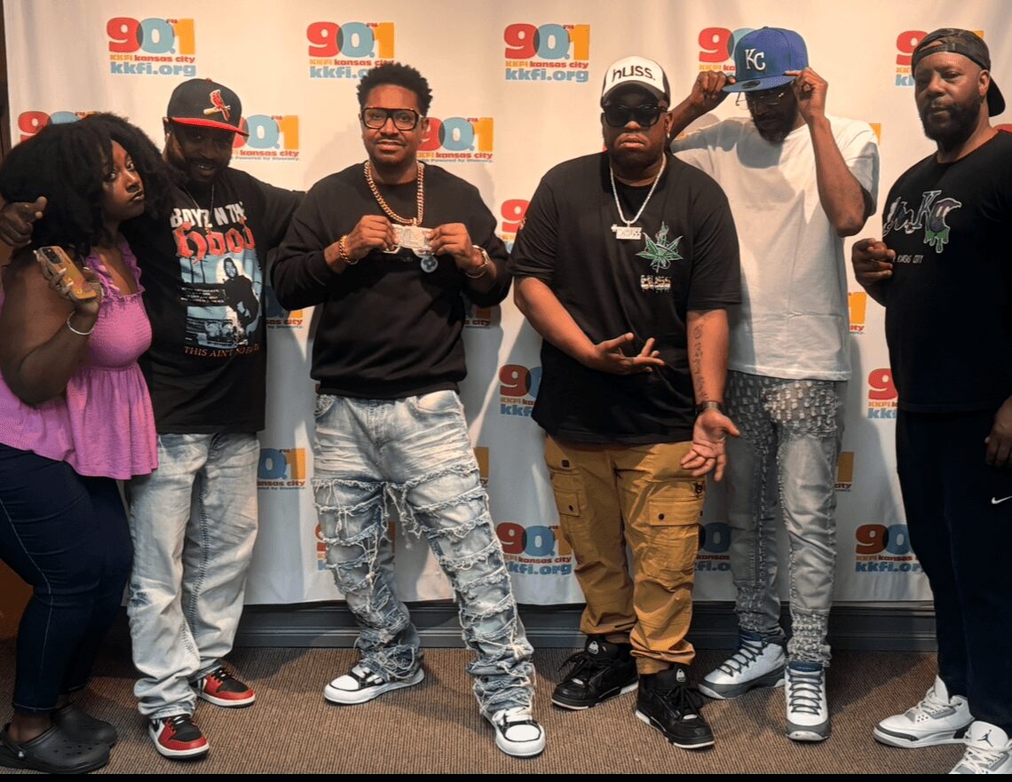 Six people standing in front of a backdrop with logos, some holding phones or making gestures, diverse in appearance.